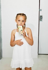 Ein Kind trägt ein weißes, ärmelloses Kleid mit einem Rüschensaum und hält einen gelben Apfelsaftkarton mit einem Trinkhalm. Ein einfacher Hintergrund mit einer Tür.
