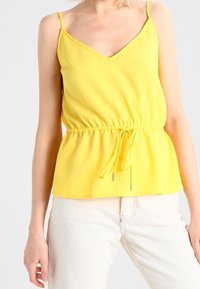 Top amarillo sin mangas con escote en V, cintura con cordón y una tela texturizada. Combinado con pantalones de color claro.
