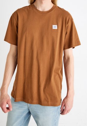 Mand iført en ensfarvet brun kortærmet t-shirt med et lille Lee-logo på brystet og lyseblå jeans.