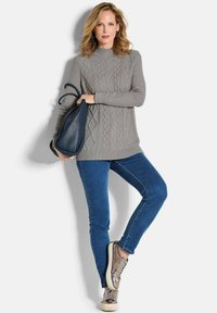 Grauer kabelgestrickter Pullover mit hohem Kragen, kombiniert mit blauen Skinny Jeans und metallischen Sneakers; hält eine marineblaue Handtasche.