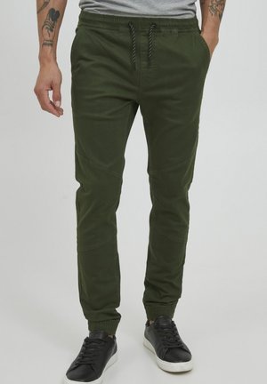 Green jogger pants made of cotton blend, featuring elastic cuffs, drawstring waist, and contrasting patches on sides. Worn with black sneakers.