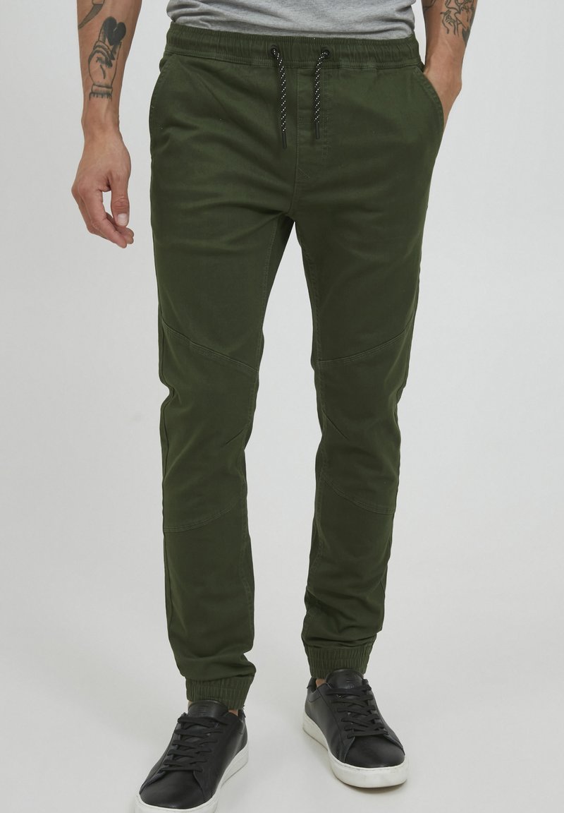 Green jogger pants made of cotton blend, featuring elastic cuffs, drawstring waist, and contrasting patches on sides. Worn with black sneakers.