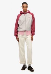 Sudadera con capucha y cremallera en gris y burdeos, con un logo, superpuesta a una camiseta a rayas rosas. Combinada con unos pantalones color crema y zapatos negros.