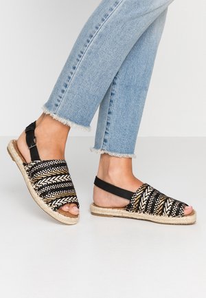 Des pieds portant des sandales à talons tressées noires, beiges et blanches, associées à un jean bleu clair avec ourlet effiloché sur un fond uni.