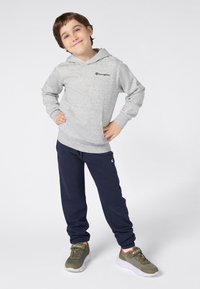 Felpa grigia con tasca frontale e pantaloni della tuta blu navy. La felpa presenta un logo sul petto e polsini a costine; le scarpe sono verdi con accenti arancioni.
