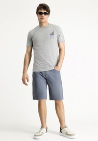 Camiseta gris de manga corta con un estampado gráfico azul en el pecho, combinada con pantalones cortos azules y zapatillas blancas con acentos verdes.