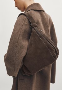 Dark brown suede bag with a rounded shape, secured with a wide strap featuring silver hardware. Worn over a textured brown coat.