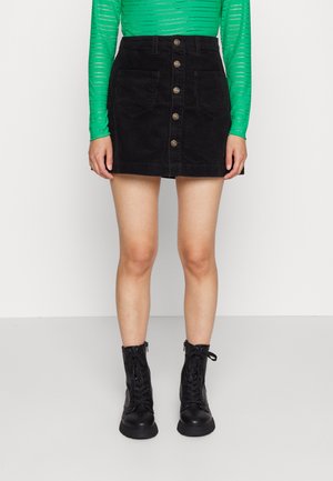Jupe en velours côtelé noir avec fermeture à boutons sur le devant, deux poches avant et coupe droite. Assortie avec un haut à manches longues vert et des bottes noires.