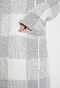 Hand teilweise in der grauen und weißen karierten Strickpullover-Tasche, die weichen Texturen des Stoffes und die Details des Bündchens zeigend.