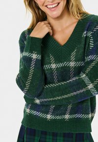 Lächelnde Person in einem grün karierten Pullover mit V-Ausschnitt und einem passenden grün-blauen karierte Rock, die mit einer Hand in der Nähe ihres Gesichts posiert.