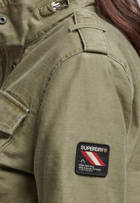 Olivgrön jacka med ett texturerat material, som har en svart lapp med röda och vita ränder märkt "SUPERDRY" på vänster ärm.