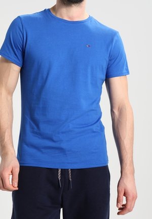 Man wearing a plain blue short-sleeve t-shirt and dark pants with a drawstring, standing against a plain light background.