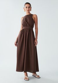 Sieviete ar savilktiem matiem, valkājot lielus zeltainus riņķus, bez piedurknēm brūnā maxi kleitā un baltās plakanās sandales uz vienkārša fona.