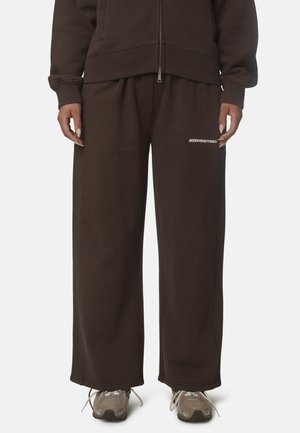 Personne portant un pantalon large marron foncé avec un petit texte blanc sur la cuisse gauche, associé à des baskets beiges.