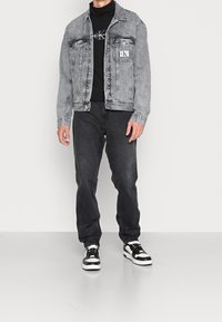 Mann trägt eine verblasste graue Jeansjacke, einen schwarzen Calvin Klein Rollkragenpullover, dunkle Jeans und schwarz-weiße Sneakers und steht auf einem grauen Boden.