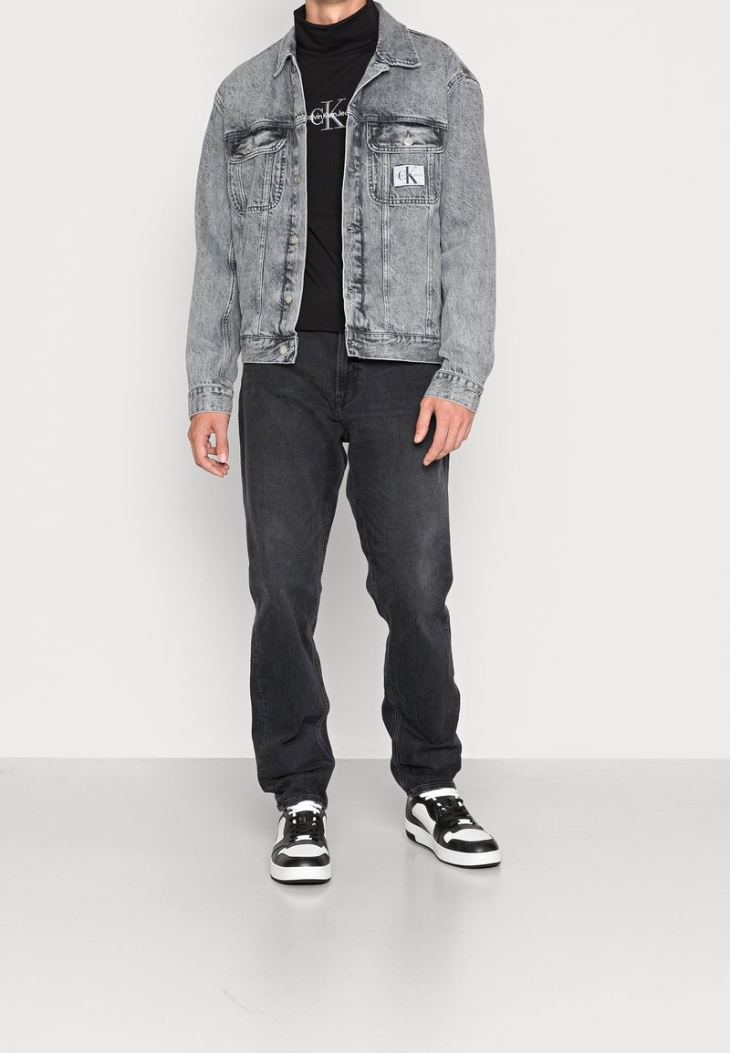 Mann trägt eine verblasste graue Jeansjacke, einen schwarzen Calvin Klein Rollkragenpullover, dunkle Jeans und schwarz-weiße Sneakers und steht auf einem grauen Boden.