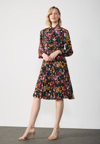 Sateen midi mekko tummalla taustalla, jossa on punaisia, vaaleanpunaisia ja keltaisia kukkia. Pitkät hihat ja poimutettu muotoilu. Yhdistetty kullanvärisiin korkokenkiin.