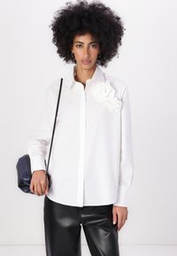 Camicia bianca con bottoni, adornata da un motivo floreale tessuto sulla spalla sinistra, abbinata a pantaloni in pelle sintetica nera e una piccola borsa scura.