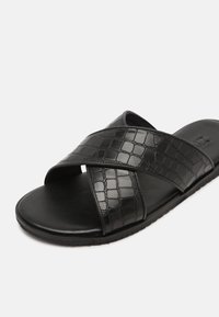 Sandale noire à enfiler avec des larges brides croisées présentant une texture crocodile et une semelle plate sur fond blanc.