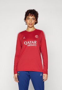 Röd långärmad tröja med vita detaljer och en logotyp. Har "QATAR AIRWAYS" i vitt, tillverkad av lätt material. Kombineras med blå byxor.