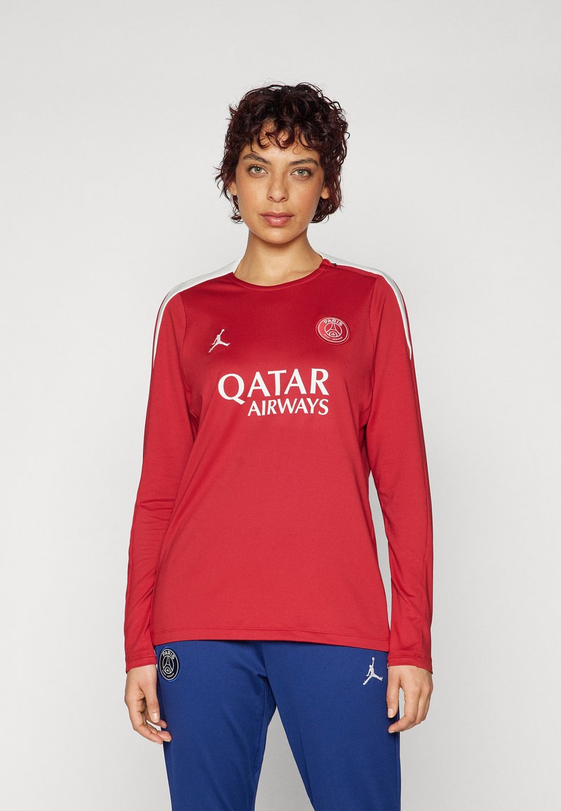 Röd långärmad tröja med vita detaljer och en logotyp. Har "QATAR AIRWAYS" i vitt, tillverkad av lätt material. Kombineras med blå byxor.