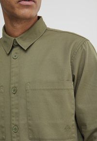 Olive green button-up shirt made from smooth fabric, featuring a classic collar, two chest pockets, and subtle logo detail on the pocket.