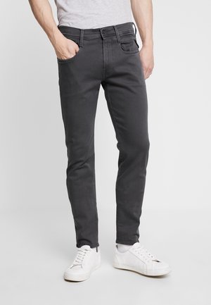 Mężczyzna ubrany w ciemnoszare slim-fit dżinsy i białe trampki, stojący z jedną ręką w kieszeni na jednolitym tle.