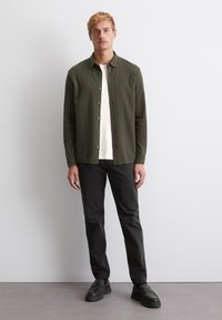 Olive green button-up shirt in soft fabric, white t-shirt underneath, paired with black pants and black slip-on shoes. Simple design and clean lines.