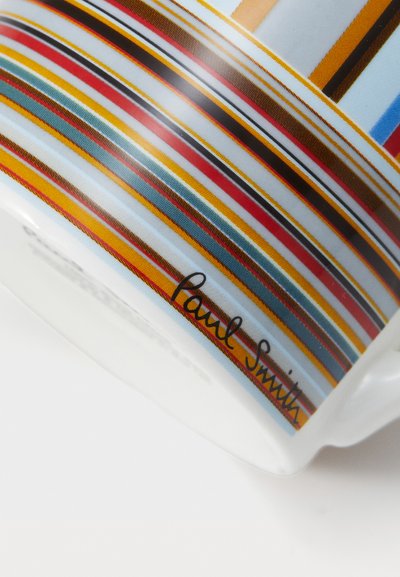 Caneca de cerâmica com riscas horizontais multicoloridas em vermelho, amarelo, azul e preto. Assinatura "Paul Smith" em itálico na superfície.