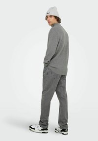 Grauer Pullover mit langen Ärmeln, kombiniert mit grauen Jeans. Weiße und schwarze Sneaker. Hellgraue Rippmütze mit einem Logo. Stehende Position.