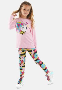 Maglietta rosa a maniche lunghe con un grafico di un'ape dei cartoni animati, abbinata a leggings floreali colorati. Sneaker bianche con lacci arcobaleno completano l'outfit.