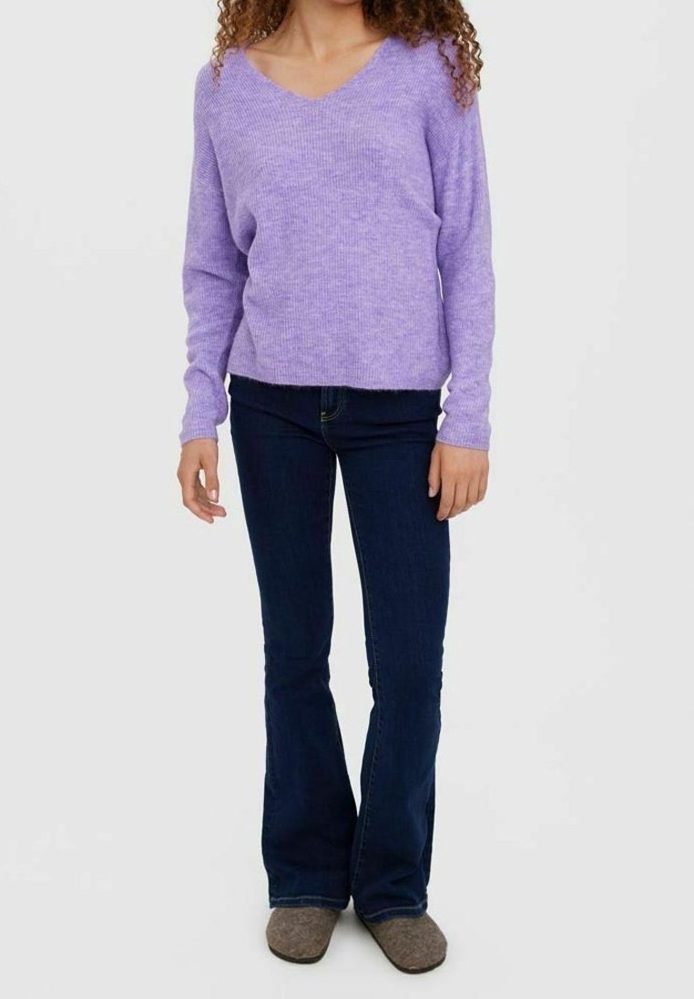 Femme portant un pull violet clair à col en V avec manches longues, un jean évasé bleu foncé et des chaussures marron, debout devant un fond uni.