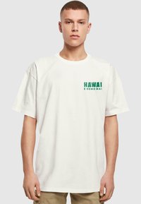 Hvit bomulls t-skjorte med grønn tekst "HAWAI" og "e komo mai" på venstre bryst, korte ermer og en avslappet passform.