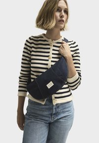 Borsa a spalla in denim con forma curva, colore blu scuro e cuciture a contrasto. Presenta un'etichetta sul davanti ed è abbinata a un maglione a righe.