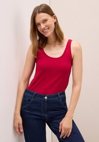 Červený top bez rukávů vyrobený z elastického materiálu, zkombinovaný s tmavomodrými džínami s knoflíkovým zapínáním a předními kapsami.