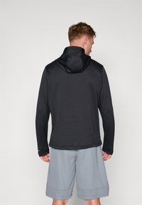 Svart hettegenser med en teksturert mønster, tettsittende hette og lange ermer, kombinert med lysegrå shorts som viser en avslappet passform.