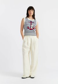 Maglione senza maniche a righe nei colori bianco, blu marino e rosso, con una grafica ad ancora; abbinato a pantaloni bianchi larghi e scarpe nere.