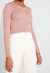 Dlouhý rukáv, žebrovaný top světle růžové barvy s kulatým výstřihem, zkrácený střih a texturovaný povrch, v kombinaci s bílými kalhotami s vysokým pasem.