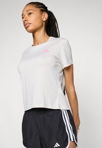 Világosszürke sportpóló rövid ujjakkal és laza szabással, pink Adidas logóval. Fekete rövidnadrággal és három fehér csíkkal viselve.