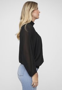 Schwarze transparente Bluse mit hohem Kragen und Rüschen-Detail. Mit langen Ärmeln und lockerem Schnitt. Kombiniert mit enganliegenden hellblauen Jeans.