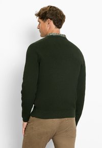Dunkelgrüner strukturierter Pullover mit einem Rippmuster und einem runden Kragen. Ergänzt durch eine gemusterte Unterlage. Kombiniert mit beigen Hosen.