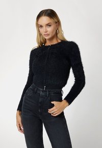 Black fluffy cardigan with long sleeves, round neckline, and front zip closure. Worn with high-waisted black jeans.