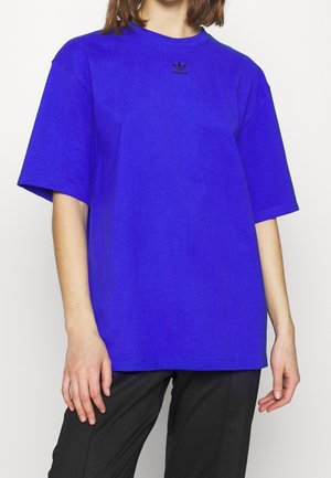 Osoba ubrana w jaskrawo niebieską oversize koszulkę Adidas z małym czarnym logo, w połączeniu z czarnymi spodniami, stojąca na białym tle.