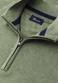 Zielony sweter z dzianiny z półzamkiem i granatowymi paskami na kołnierzu, z widoczną metką marki wewnątrz kołnierza.