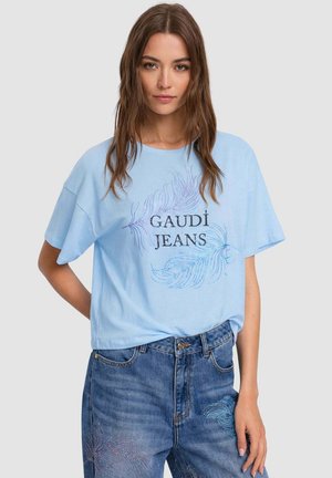 Donna con capelli castani lunghi che indossa una maglietta azzurro chiaro con la scritta "GAUDÍ JEANS" e jeans blu con delicati motivi decorativi.