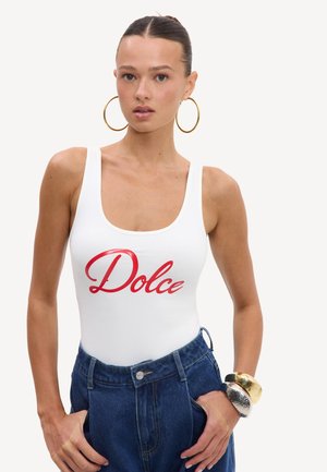 Femme avec les cheveux en chignon porte de grandes boucles d'oreilles créoles, un débardeur blanc avec un texte rouge ""Dolce"", un jean bleu taille haute et des bracelets au poignet droit.