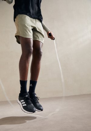 Chaussures de sport noires avec des accents blancs, shorts kaki et chaussettes bleu marine. Corde à sauter en mouvement ; mise au point sur les jambes en position de saut.