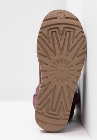 Semelle d'une botte UGG marron pressée contre un mur blanc d'angle, affichant un motif de bande de roulement détaillé avec le logo UGG au centre.