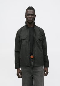 Black lightweight jacket with a zip front and stand collar, featuring two chest pockets and long sleeves, paired with dark denim jeans.