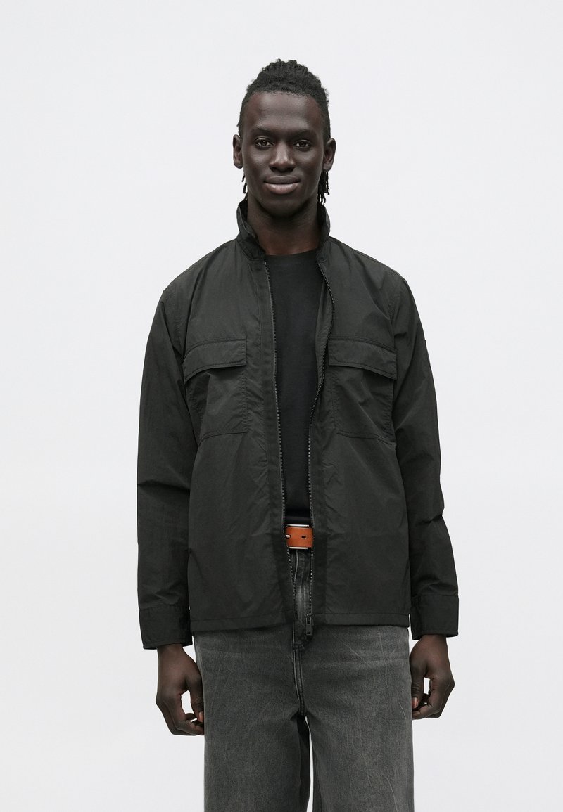 Black lightweight jacket with a zip front and stand collar, featuring two chest pockets and long sleeves, paired with dark denim jeans.